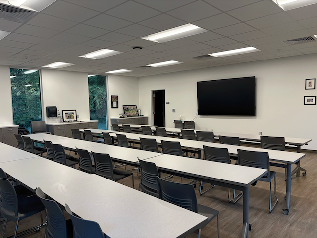 A classroom with four rows of tables and chairs facing a wall with a video screen.