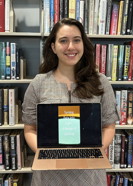 Raquel Beatriz Caldas Laranjeira holding a laptop with her Libraries Undergraduate Research Award-winning project on it, in front of books