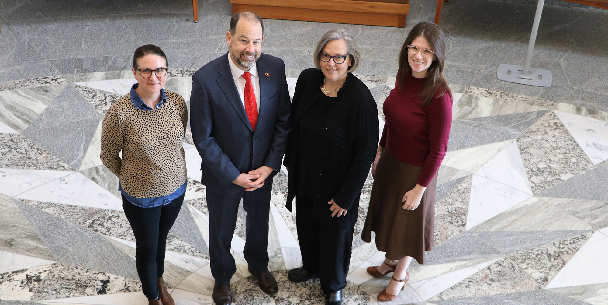 Photo of, left to right, Sara Wright, Toby Graham, Emily Gore, and Kat Stein, all smiling and looking upwards towards the camera in the Special Collections Building lobby