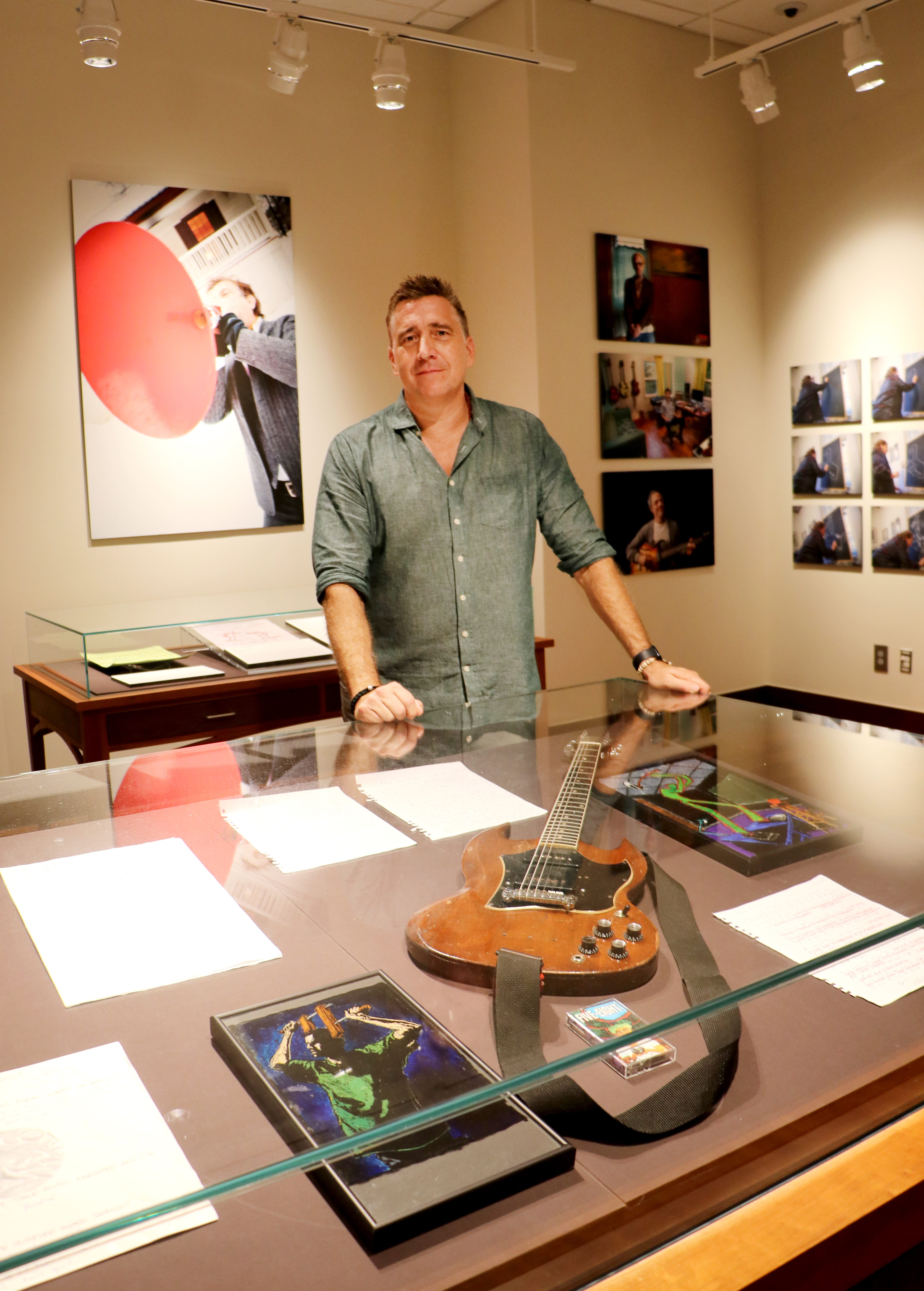 Man in blue shirt stands behind an exhibit case with a guitar and art and in front of photography