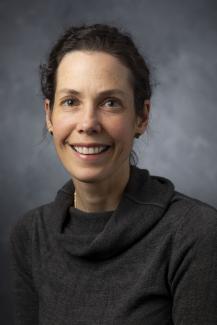 Woman with dark curly hair pulled back from her face. She's wearing a charcoal grey cowl neck sweater.
