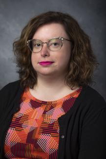 Woman with shoulder length brown curly hair. She's wearing a brightly colored patterned shirt in shades of orange, pink, and red.