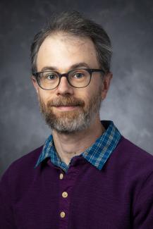 Bearded man with glasses wearing a blue shirt under a purple buttoned-up cardigan.