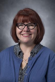 Auburn haired woman wearing a floral shirt under a blue cardigan.