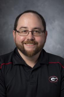 Bearded man with glasses wearing a black golf shirt with the the UGA "Big G" logo on it.