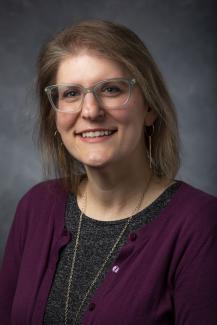 Blonde woman in glasses wearing a dark grey shirt under a magenta cardigan