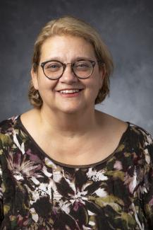 Woman with chin length blond hair wearing glasses and a floral patterned shirt.