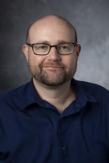 Man with a beard and glasses wearing a blue button up shirt. 