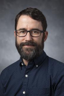Man with a beard and glasses wearing a patterned blue button down shirt.