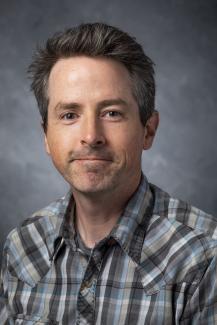 Man with brown hair wearing a plaid button up shirt.