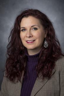 Woman with long dark red hair wearing a purple sweater under a tan blazer.
