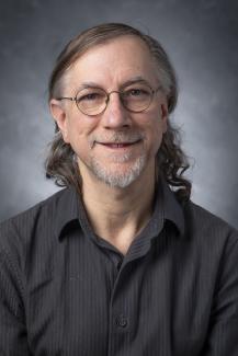 Man with curly shoulder length hair wearing glasses and dark gray button up shirt.