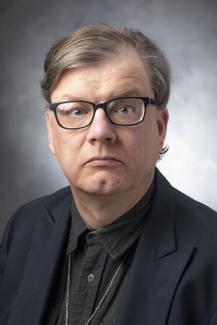Man with short light brown hair wearing glasses, a green button up shirt, and a black jacket.