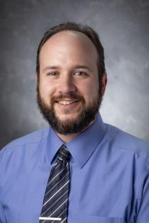 Man with dark hair and beard wearing a blue button up shirt and diagonally striped tie.