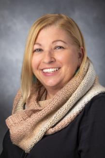 Woman with blonde hair smiling, wearing a camel scarf and dark shirt.