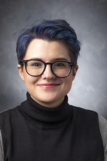Person with short purple-ish hair wearing glasses and a dark turtleneck and smiling.