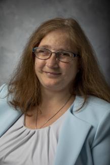 Woman with shoulder length dark red hair. She is wearing blue glasses and a light blue blaser with a white blouse.