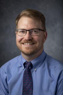 smiling man with strawberry blond hair, glasses, and a blue shirt with tie.