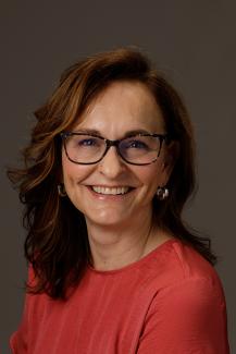 woman with coral shirt and glasses, smiling with dark hair below shoulders.
