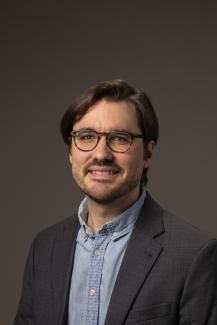 smiling man with glasses, dark hair, blue collared shirt, and dark jacket