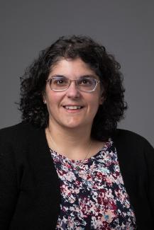Woman with dark curly hair and glasses wearing a floral shirt and black cardigan.