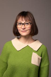 Woman with glasses and bangs wearing a green shirt with a cream collar and front pocket.