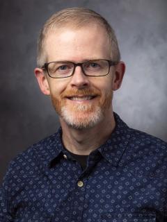 Man with pale red hair wearing glasses and a dark blue shirt.