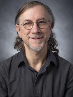 Man with curly shoulder length hair wearing glasses and dark gray button up shirt.