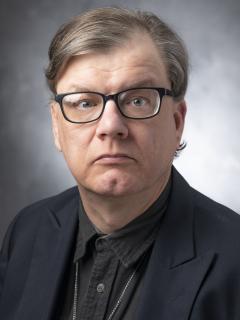 Man with short light brown hair wearing glasses, a green button up shirt, and a black jacket.