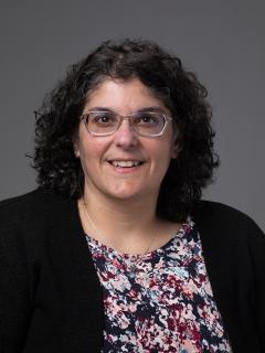 Woman with dark curly hair and glasses wearing a floral shirt and black cardigan.