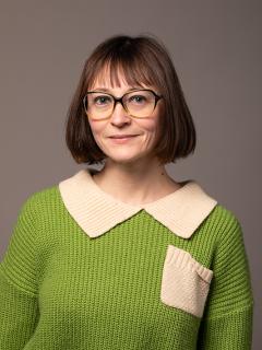 Woman with glasses and bangs wearing a green shirt with a cream collar and front pocket.