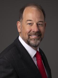 Man with short dark hair and a beard wearing a dark suit, white shirt, and red tie.