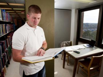 A young man consults a book, standing next to a small study table by a window with a view of campus